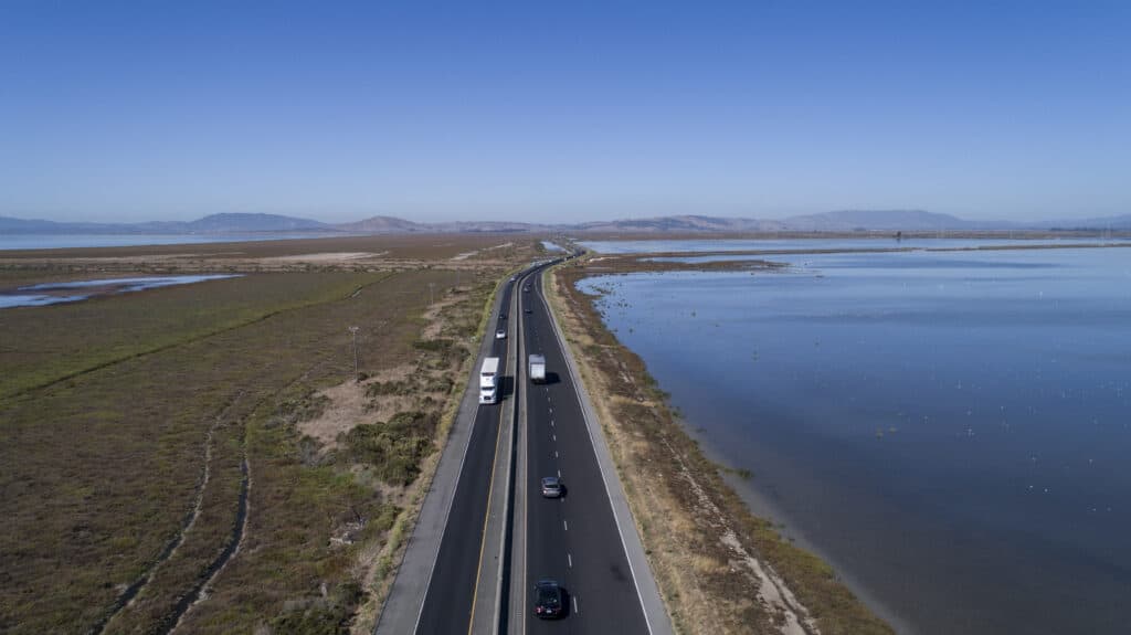 Aerial of a 3 lane highway with marsh on the left side and water on the right